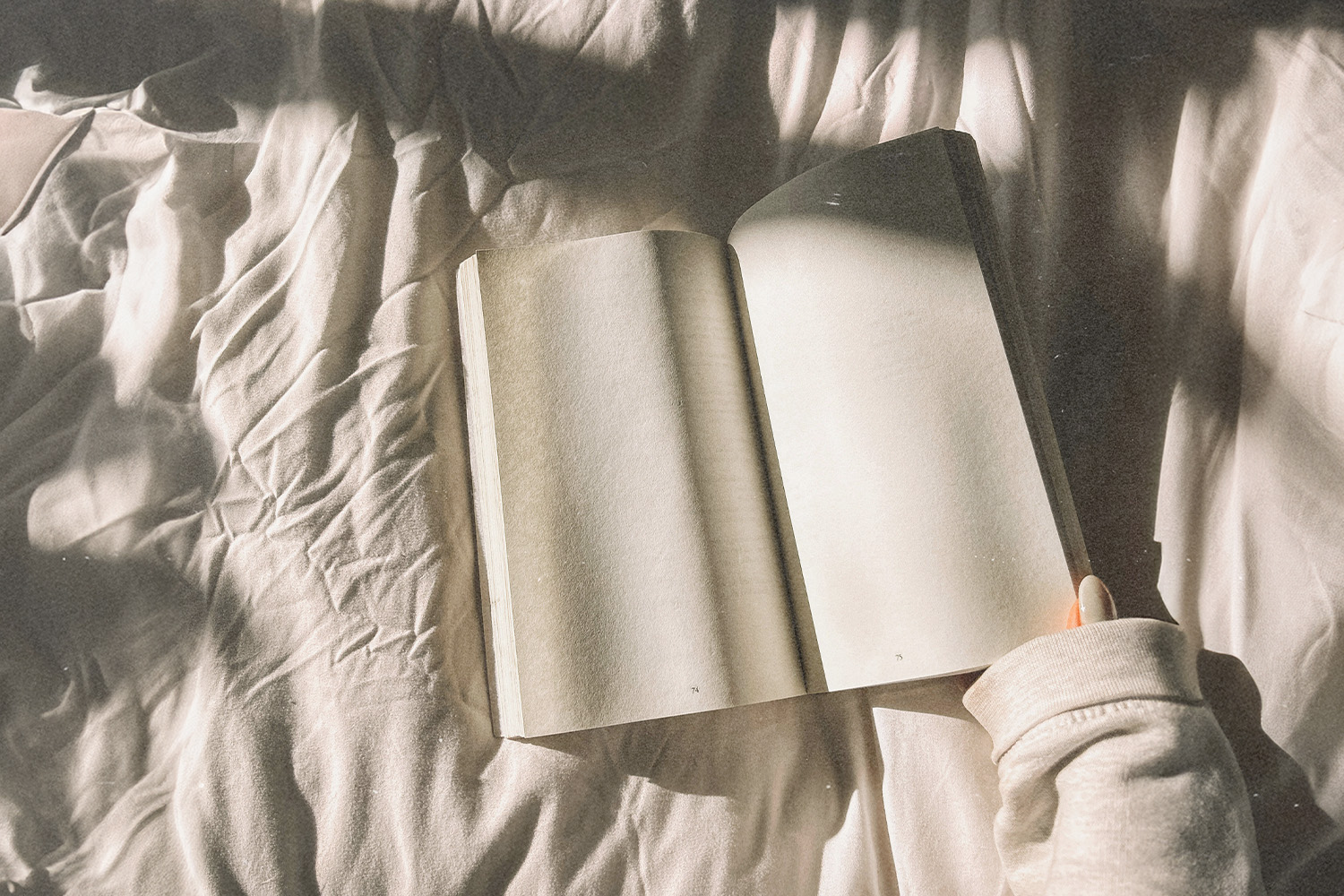 An Open Book is laid on a bed with a hand about to turn to the next page. 