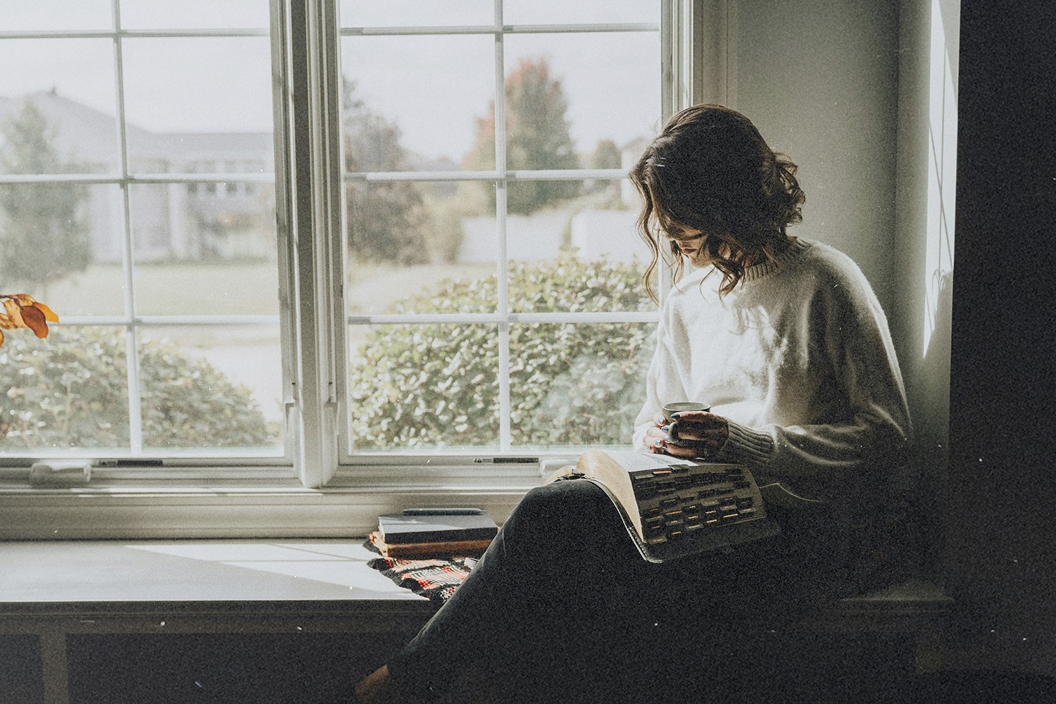 Person sitting on a windowsill with an open book and smartphone, next to stacked books and a blanket, with a large window showing greenery outside.